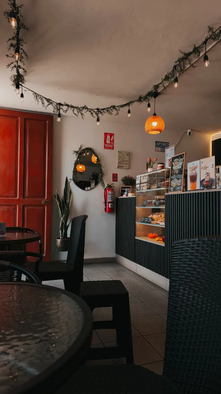 Interior de Yummy: guirnalda de luces entre plantas, lámpara de ratán sobre la barra con vitrina de postres, espejo junto a la puerta roja y mesas de mimbre